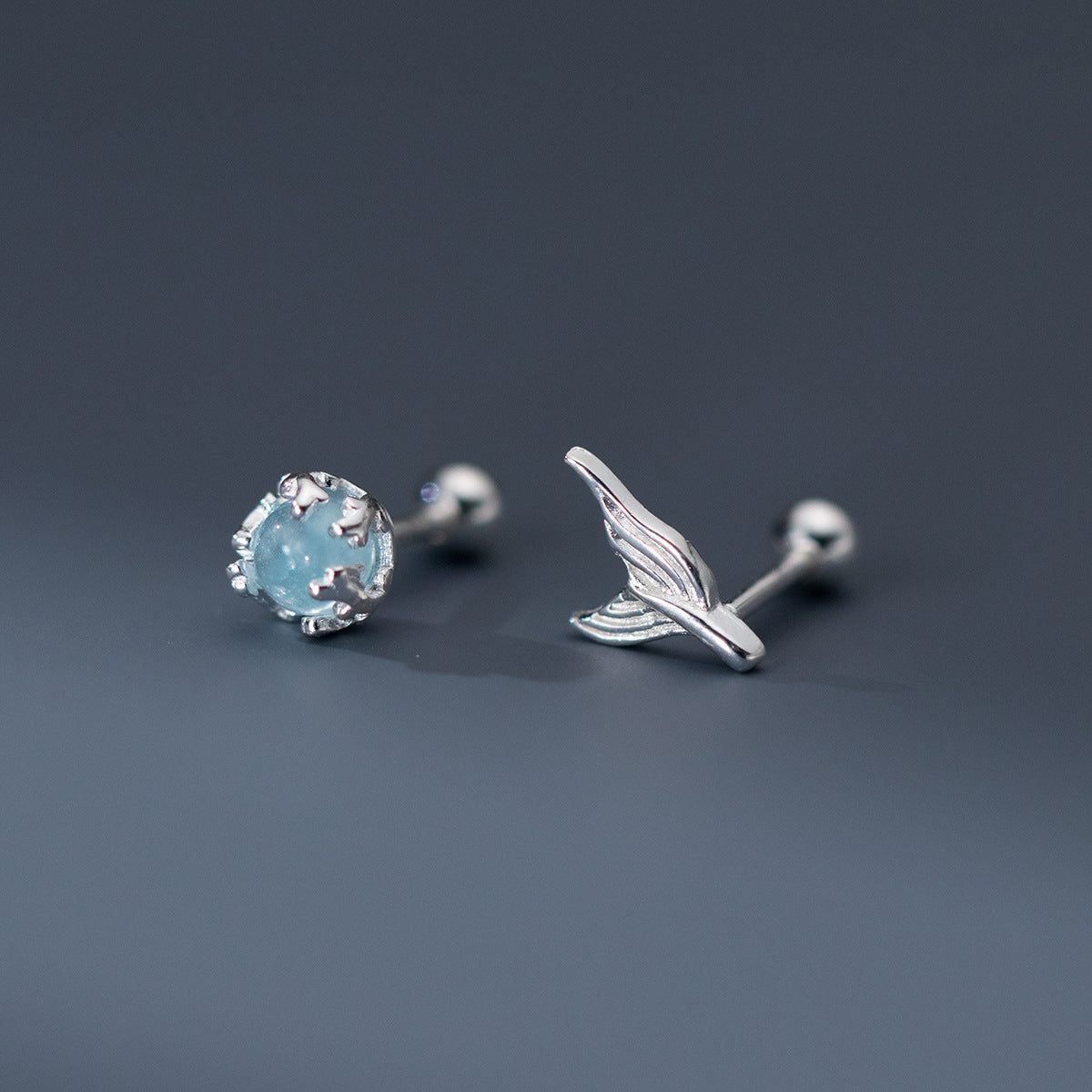A pair of 925 sterling silver mermaid tail stud earrings, showcasing the asymmetrical design with a light blue crystal stud and a separate spiral whale tail backing.