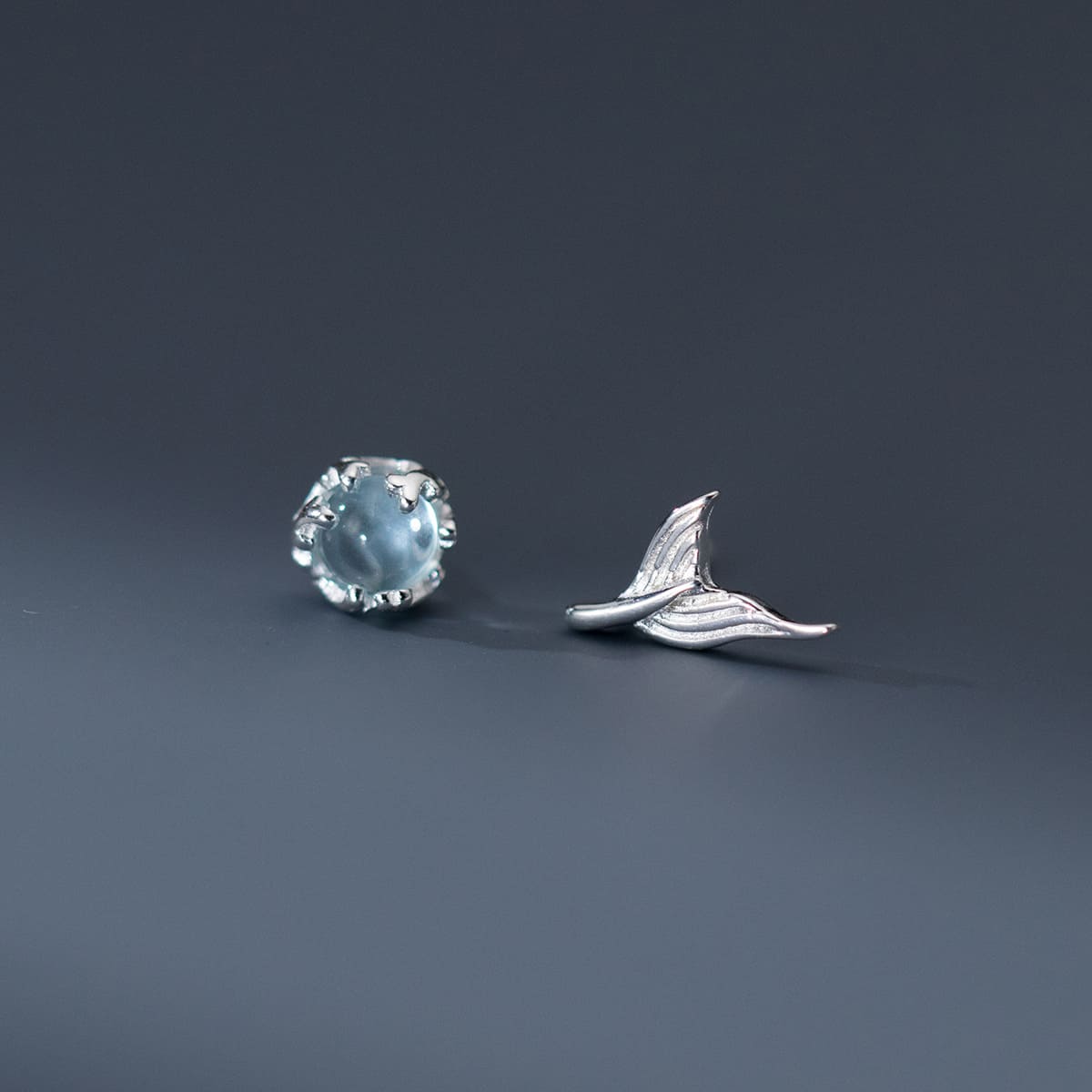 A pair of 925 sterling silver asymmetrical stud earrings, one featuring a whimsical mermaid tail design and the other a sparkling light blue gemstone.