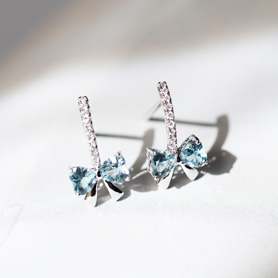 A pair of dainty 925 sterling silver bowknot stud earrings, each featuring a central light blue cubic zirconia gemstone and accented with clear crystals.