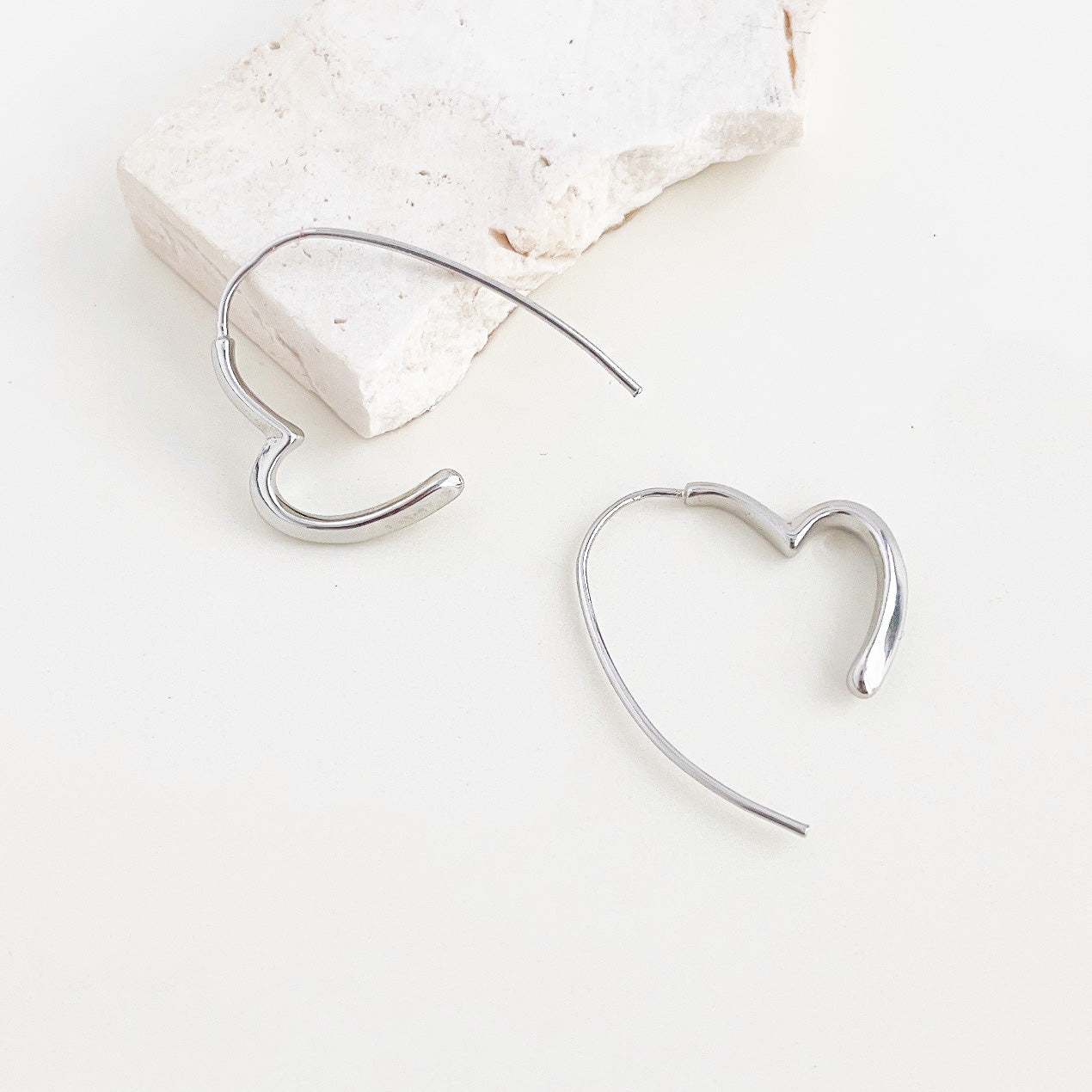 A pair of metal silver color heart line hoop earrings with a smooth, polished finish, displayed on a clean white background.