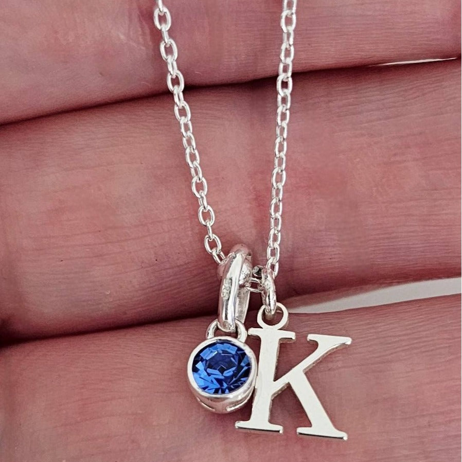 A close-up of the personalized alphabet letter necklace, showing a 'K' initial pendant in 14K gold-plated stainless steel with a vibrant blue birthstone accent.