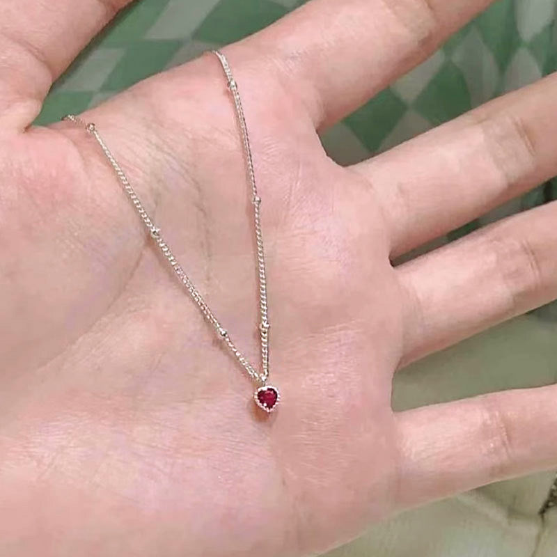 A close-up of a 925 sterling silver necklace featuring a delicate, heart-shaped red zircon pendant, showcasing its fine chain and brilliant sparkle.
