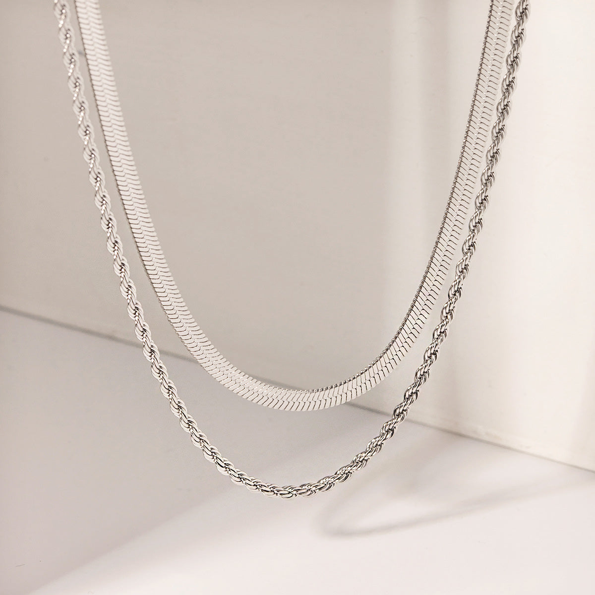 A set of two stainless steel necklaces, one with a flat herringbone chain and the other with a twisted rope design, displayed on a neutral background.