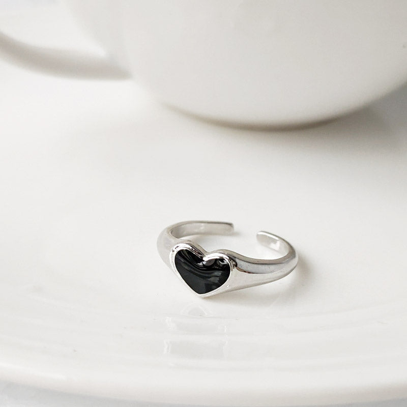 Close-up of a silver-plated ring with a black enamel heart, part of a five-piece set, displayed on a white saucer.