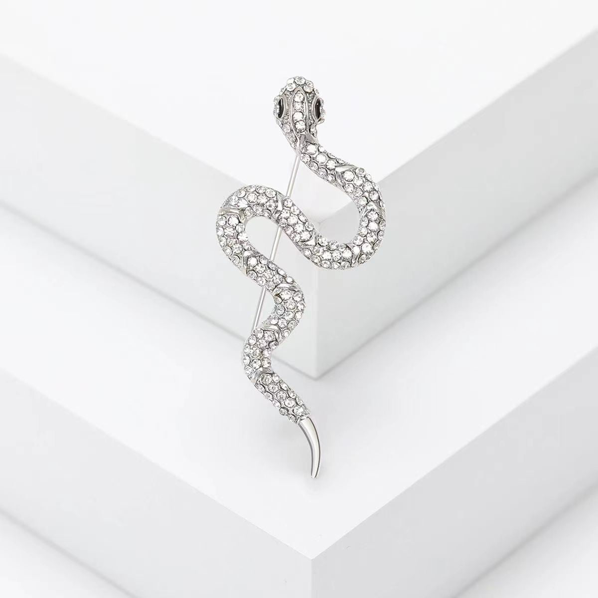 A close-up of the silver, snake-shaped brooch, intricately encrusted with sparkling clear gemstones, displayed on a white surface.