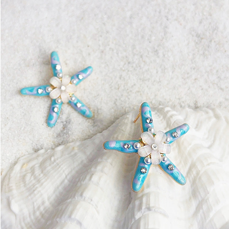 A pair of handmade enamel starfish stud earrings with blue and white details, featuring a central pearl and resin flower, showcasing a beach-inspired design.
