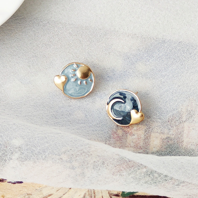 A pair of 14k gold-plated stud earrings with blue enamel, featuring an asymmetrical design of a sun and a wave, each with a small heart accent.