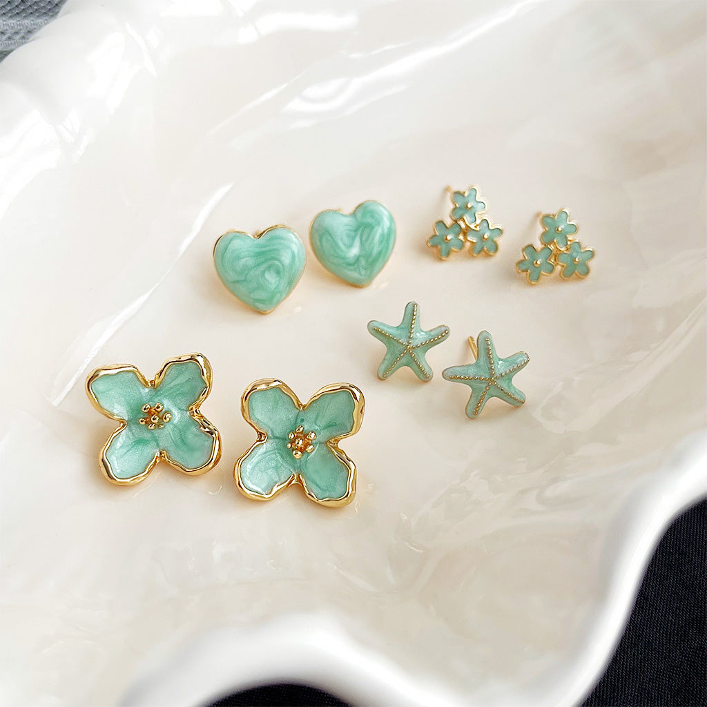 A collection of five pairs of 14k gold-plated stud earrings with mint-green enamel, featuring flower, heart, and starfish designs.