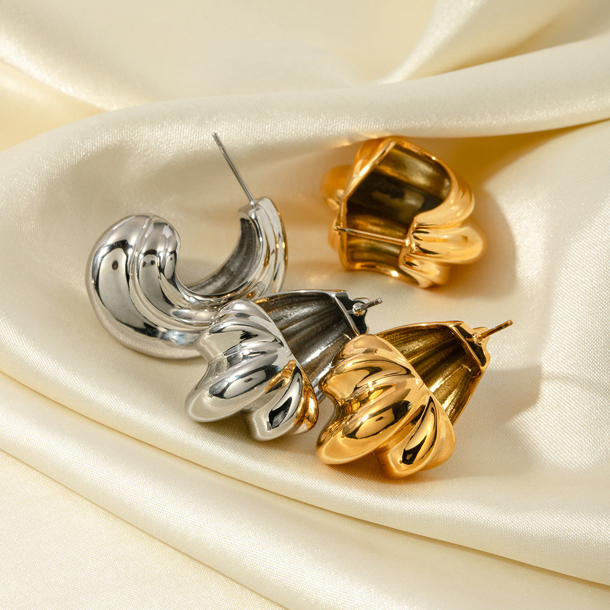 A set of stainless steel chunky geometric C-shaped hoop earrings in both gold and silver finishes, displayed elegantly on a cream-colored satin background.