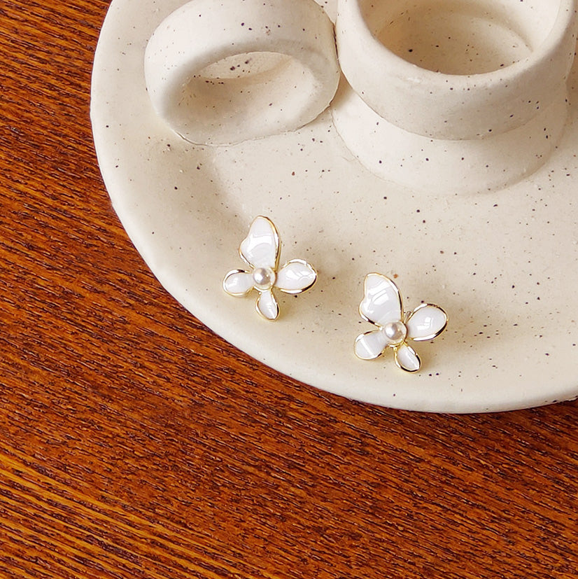 A pair of small white enamel butterfly stud earrings with gold trim and a central pearl, displayed on a decorative ceramic plate.