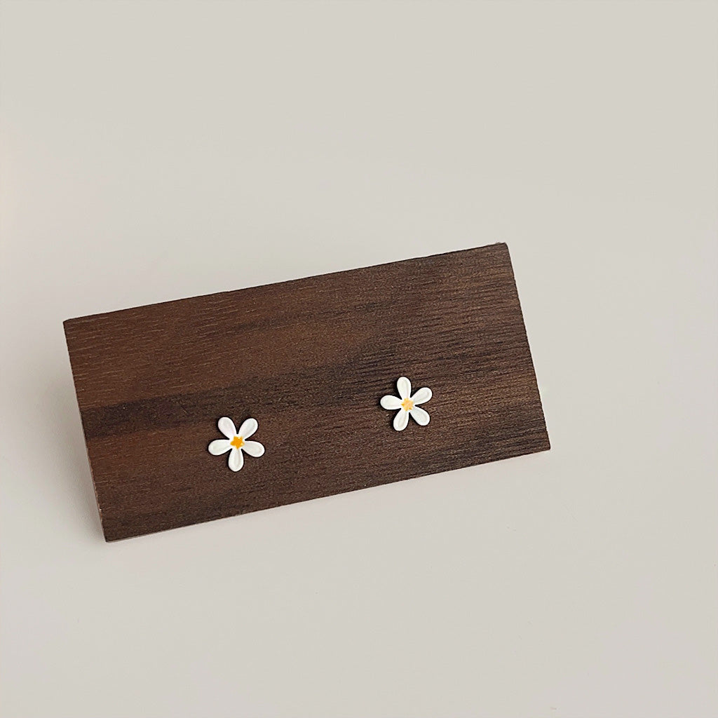 A pair of wholesale cute mini enamel daisy flower stud earrings displayed on a rustic wooden block, highlighting their white petals and yellow centers.