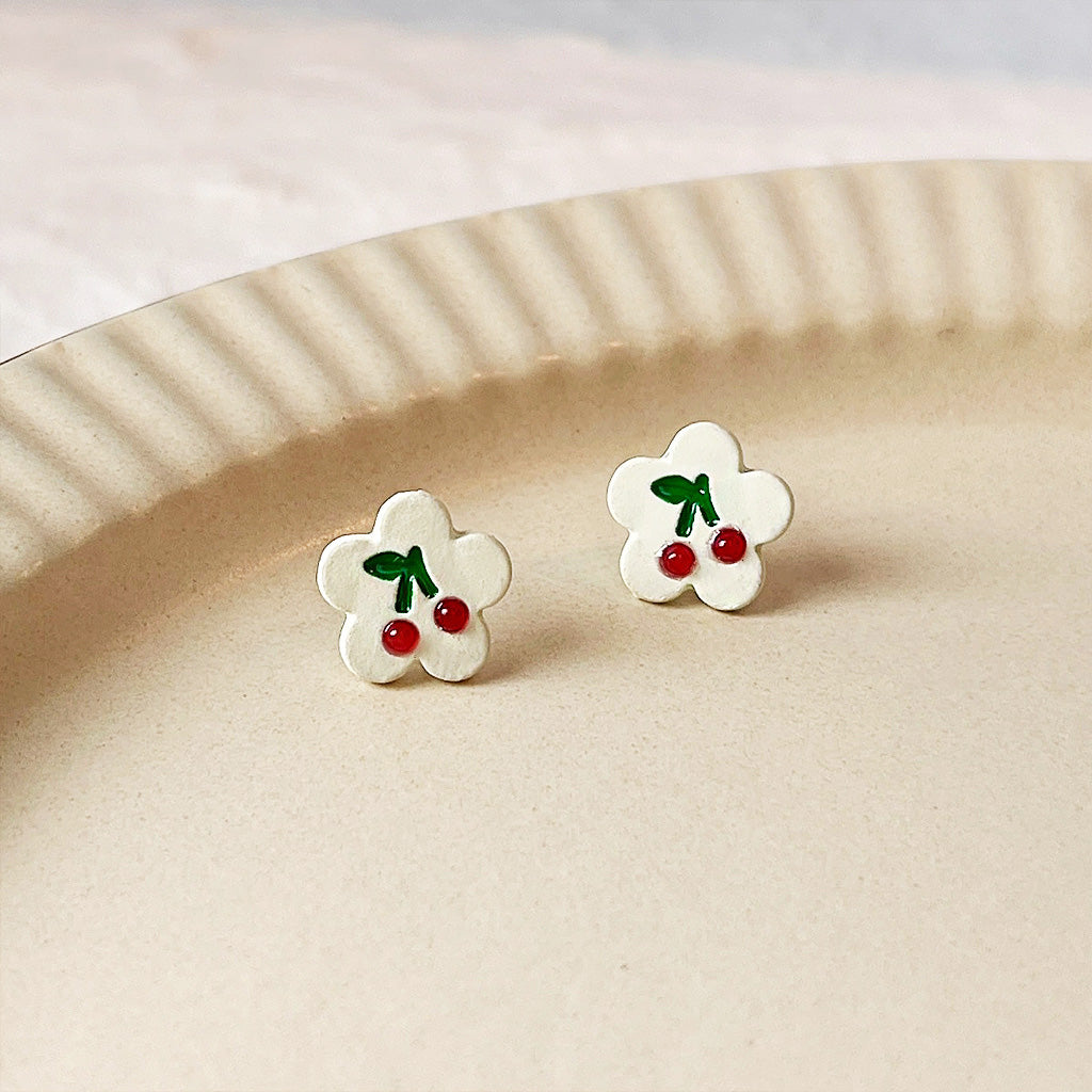 A pair of cute white flower-shaped stud earrings with a vibrant red enamel cherry pattern, crafted from brass by JR Fashion Accessories.