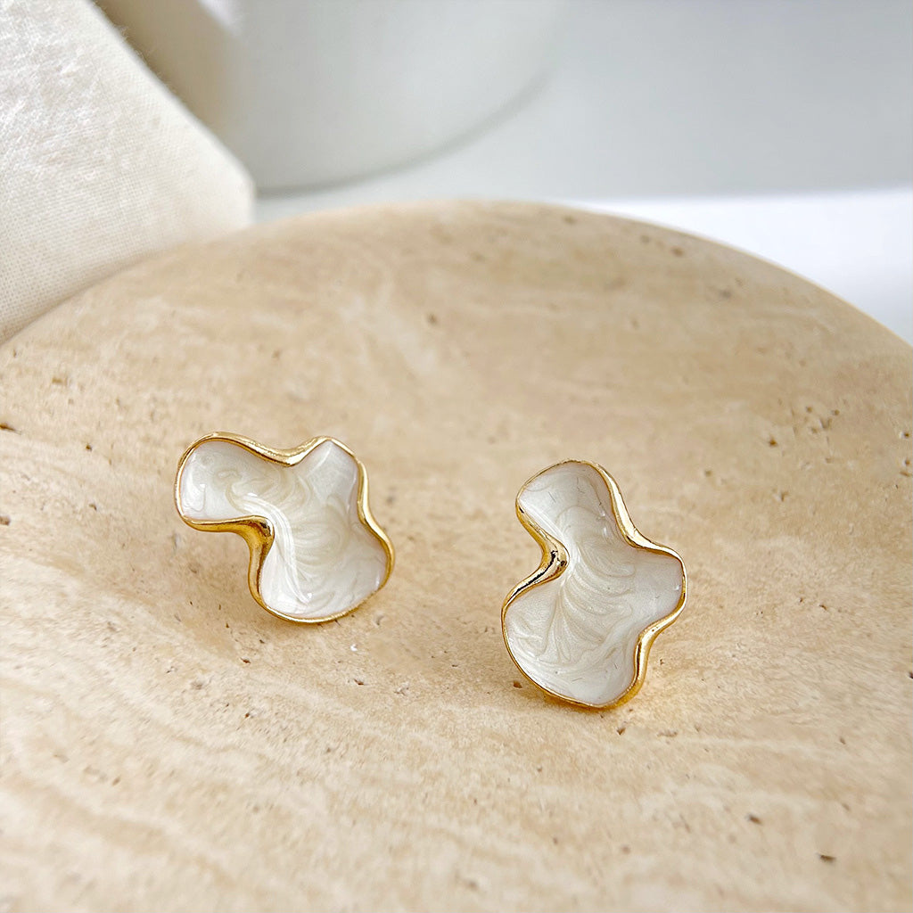 A pair of 14k gold plated stud earrings with a unique, irregular shape and white enamel inlay, displayed on a neutral background.
