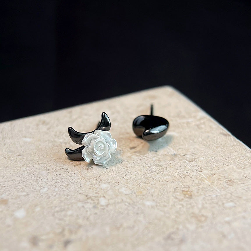 A pair of handmade asymmetrical stud earrings, one featuring a white resin rose on a black base and the other a simple black stud, showcasing the unique floral design.