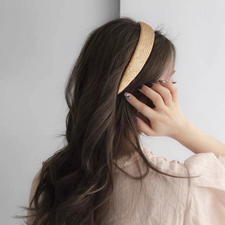A woman with long dark hair modeling a stylish wide beige straw woven braided headband, showcasing its bohemian-chic look.