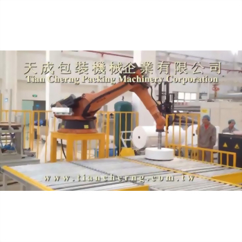不織布包裝設備 18-2 - 工廠內橘色機械手臂正將白色圓柱形物品精準地放置在黃色滾輪輸送帶上,背景可見廠房與人員。
