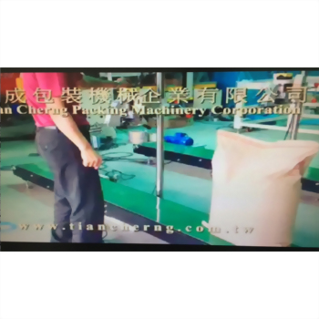 紙袋縫袋機 11-1 - 廠房內,一名穿紅色上衣的員工站在綠色輸送帶旁,一個淺粉色大包裝袋立於地面,背景可見天成包裝機械企業的文字招牌。